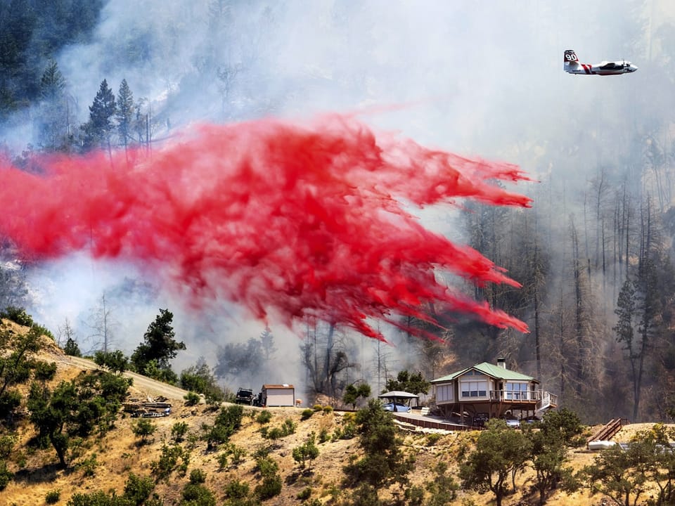 Flugzeug verstreut roten Feuerlöschmittel über brennende Wälder und Häuser.