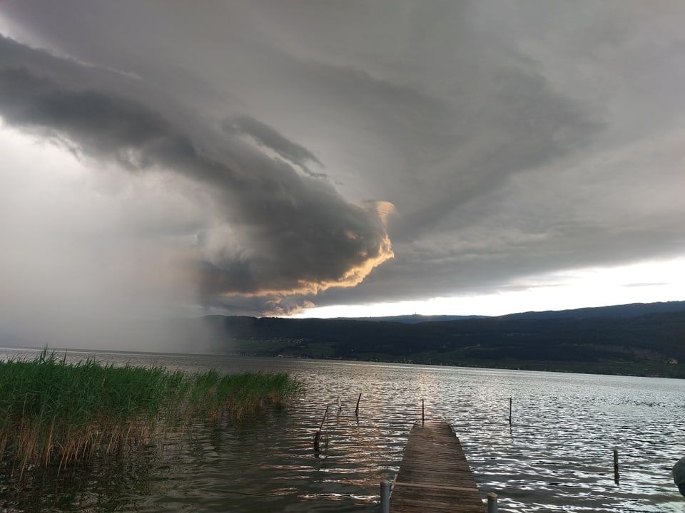 Dunkle Wolken über einem See mit Steg und Schilf im Vordergrund.