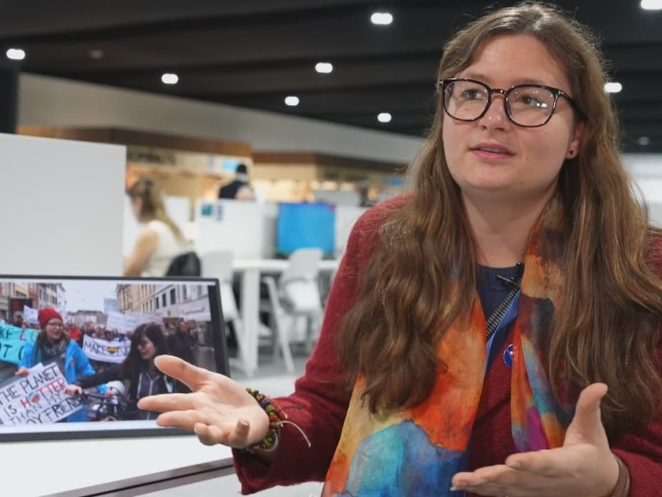 Frau mit Brille spricht, im Hintergrund Protestbild.