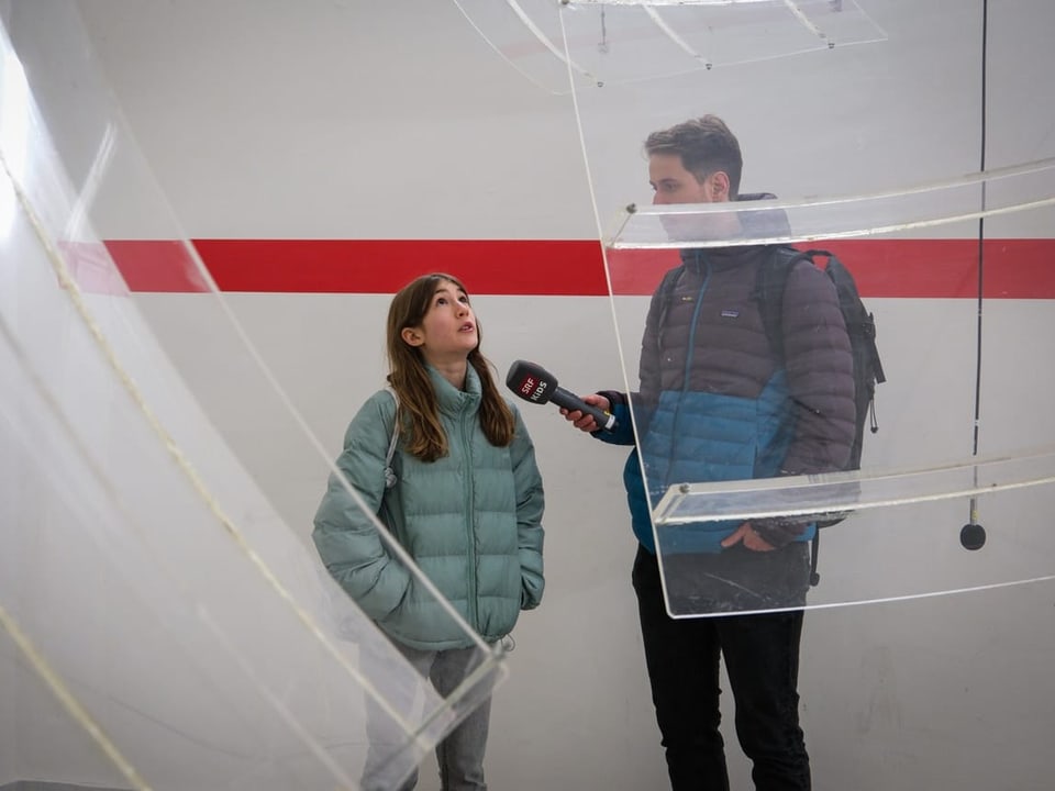 Zwei Menschen stehen zwischen Plastikscheiben, die von der Decke hängen. 