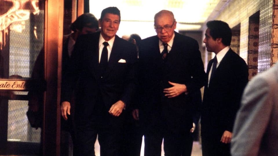 Zwei Männer in schwarzen Anzügen stehen vor einem Eingang. Rechts davon auch ein Mann in dunklem Anzug. 