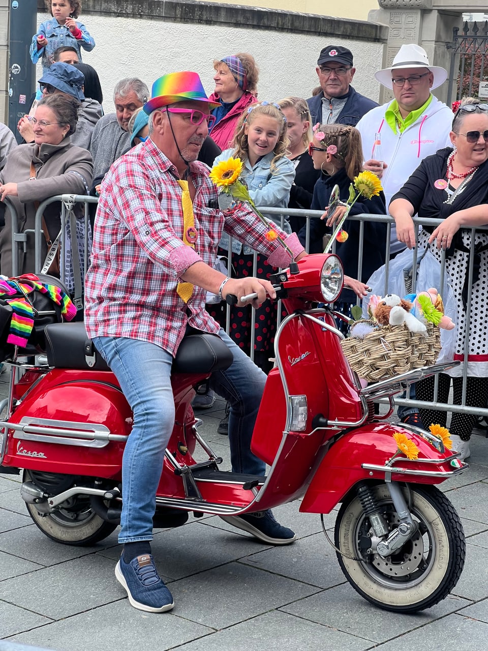 Mann mit bunter Mütze und Sonnenbrille auf rotem Roller während einer Parade.