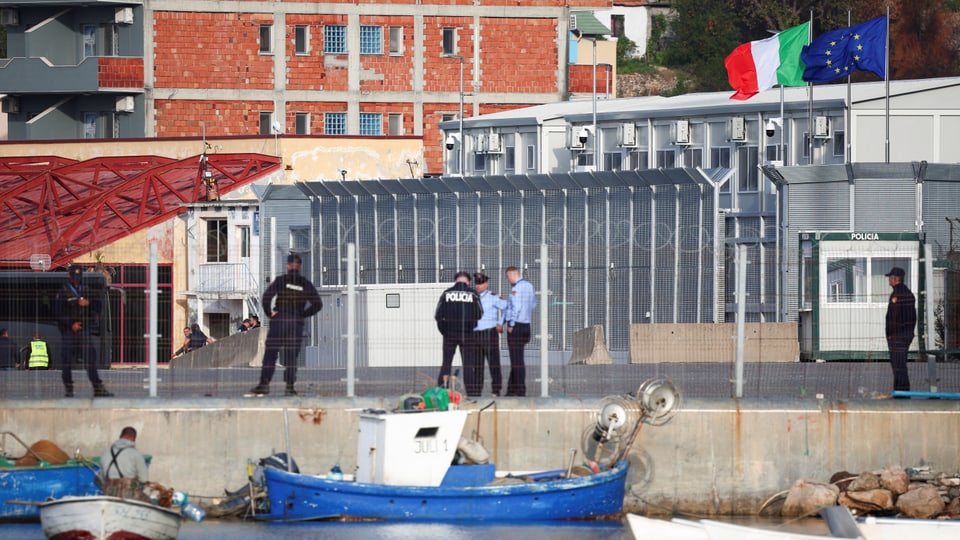 Polizisten stehen hinter einem Zaun vor einer Gebäudeanlage, mit Booten im Vordergrund.