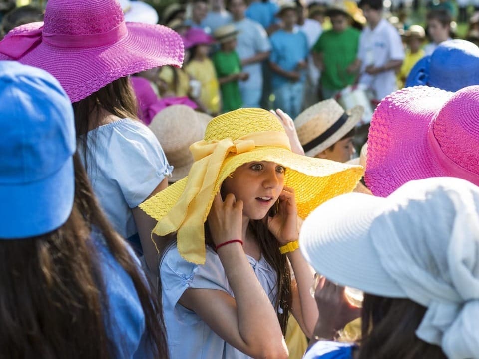 Ein Mädchen mit gelbem Sonnenhut hält sich die Ohren zu. Daneben viele andere Kinder mit bunten Hüten. 