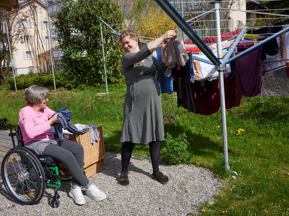 Ältere Frau im Rollstuhl gibt Kleidung an eine jüngere Frau weiter, die Wäsche aufhängt.