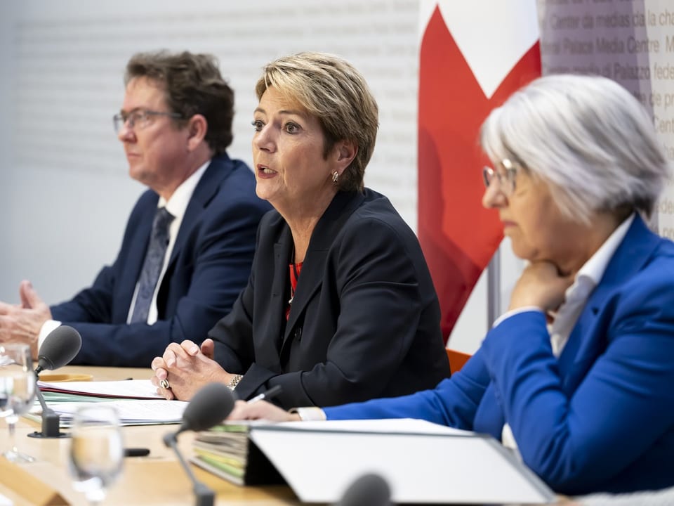 Drei Personen sprechen bei einer Konferenz, Schweiz-Fahne im Hintergrund.