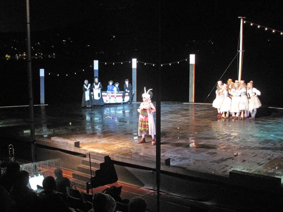 Eine Freilicht-Theaterbühne in der Nacht. Im Vordergrund eine Einzelperson, im Hintergrund zwei Theatergruppen.