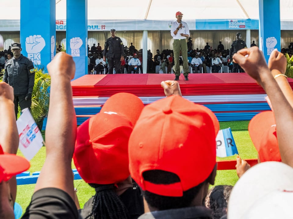 Person hält Rede auf roter Bühne vor Publikum mit roten Hüten.