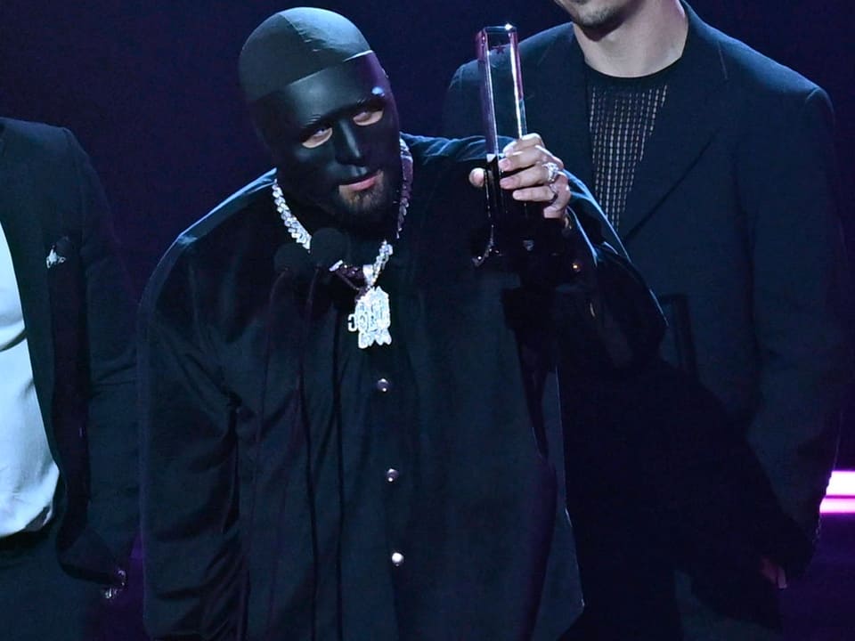 Person in schwarzer Maske hält eine Trophäe bei einer Veranstaltung.