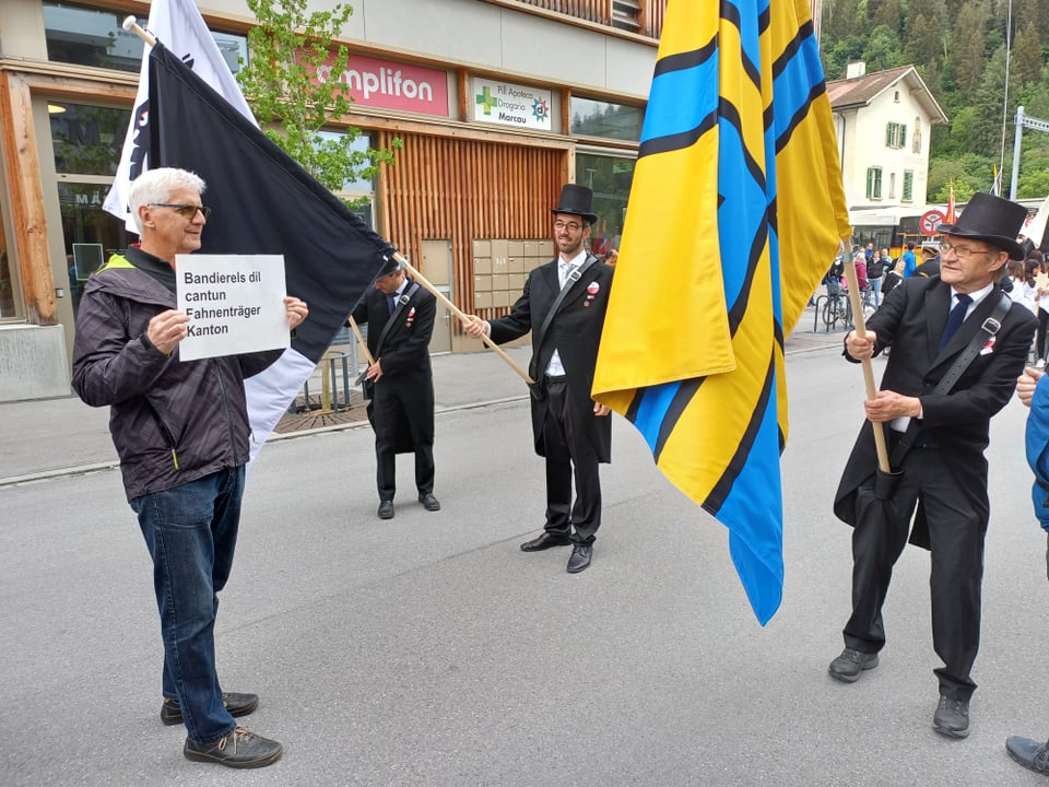 Mann hält Kantonsfahne bei Parade, umgeben von Männern in traditionellen schwarzen Anzügen.