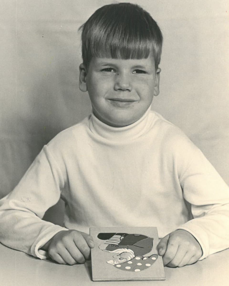 Ein Bub mit einem weissen Rollkragenpullover sitzt vor einem Puzzle aus Holz.