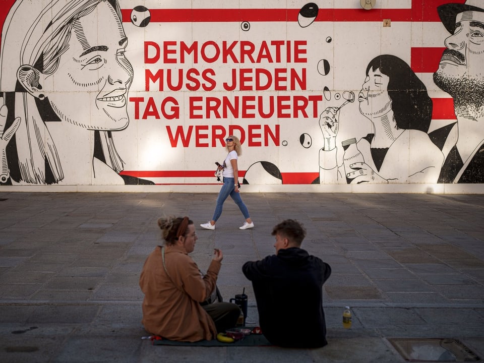 Strassenszene vor Wandgemälde mit Schriftzug 'Demokratie muss jeden Tag erneuert werden'.