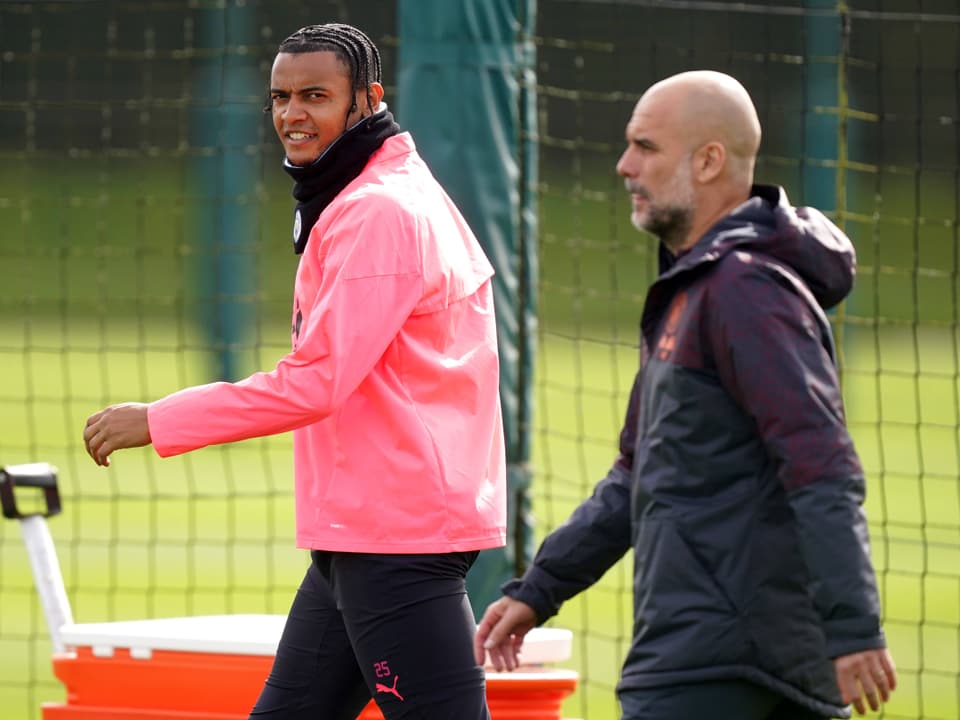 Zwei Männer auf einem Fussballplatz, einer in einem pinken Trainingsanzug, der andere in einem schwarzen Mantel.