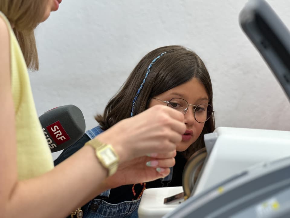 Eine Frau hält etwas vor einen Schleifstein. Ein Kind schaut nebendran neugierig zu.