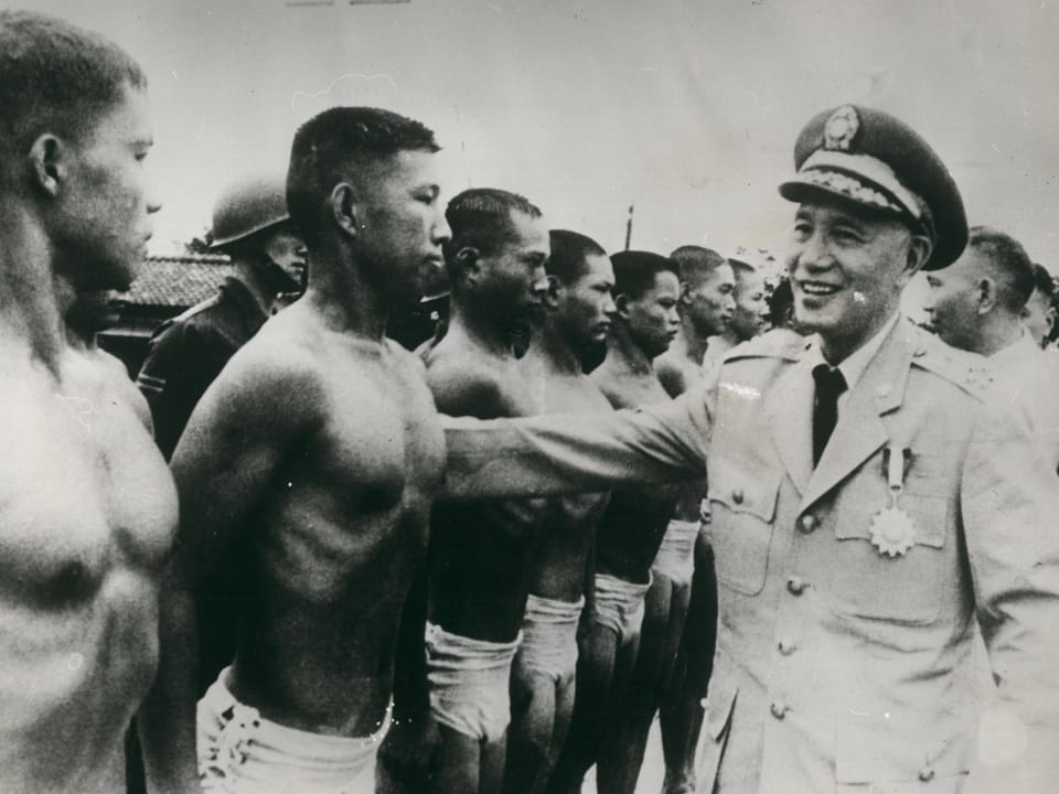 Ein Mann in Militäruniform schreitet eine Reihe von trainierten Männern ab, die nur in kurzen Hosen bekleidet sind.