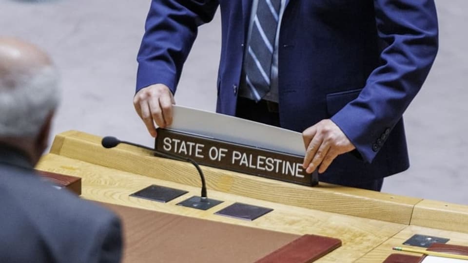 Person stellt Schild mit Aufschrift 'State of Palestine' auf einem Tisch auf.