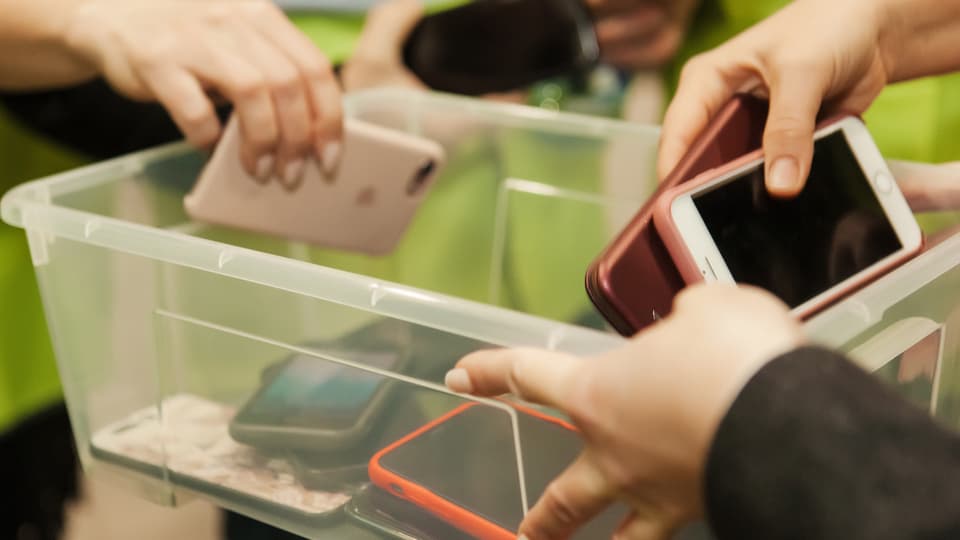 Hände legen Smartphones in einen durchsichtigen Behälter. 