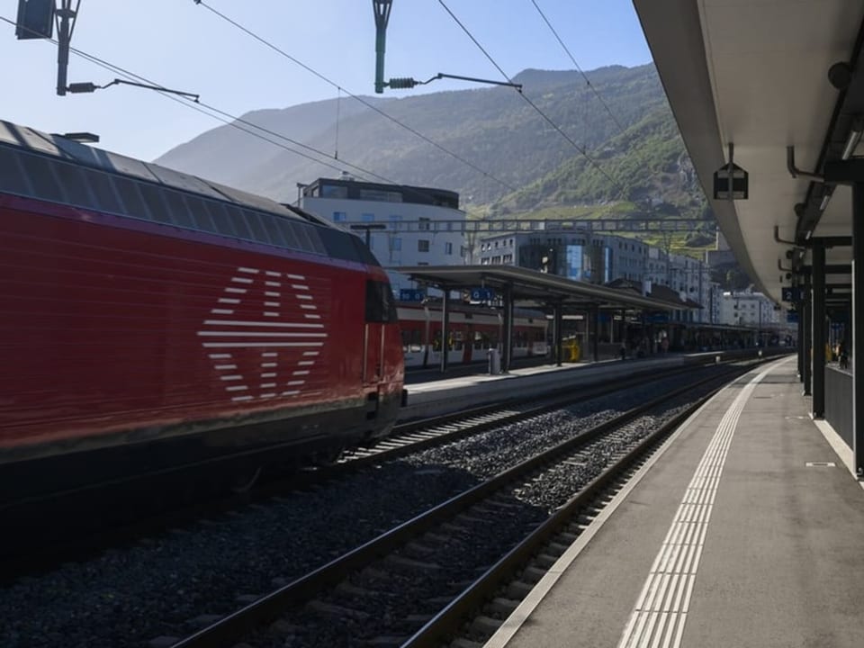 Bahnhofsszene mit Zug und Berge im Hintergrund.