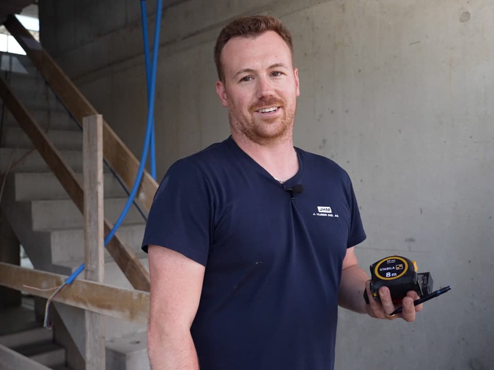 Mark Huber lächelt in die Kamera. Er ist auf einer Baustelle und hält Massband, Stift und Wasserwaage in der Hand.