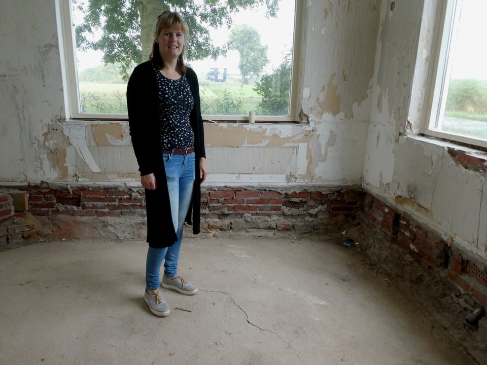 Ein Frau steht in einem Wohnzimmer, die Farbe von den Wänden abgeblättert, ein Riss im Boden.