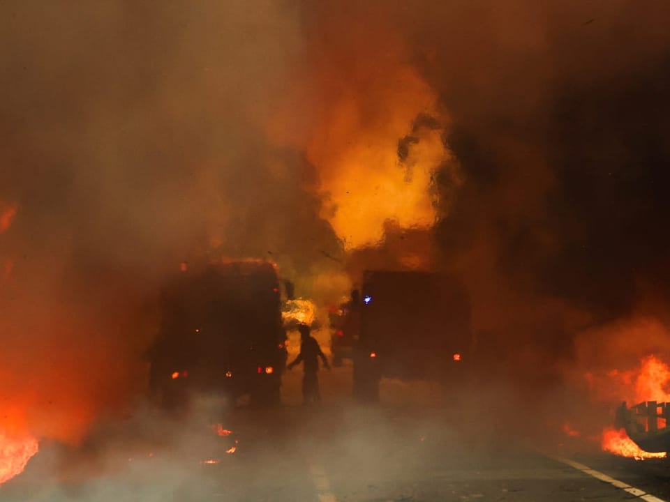 Silhouette einer Person und zwei Fahrzeuge inmitten von Flammen und Rauch.