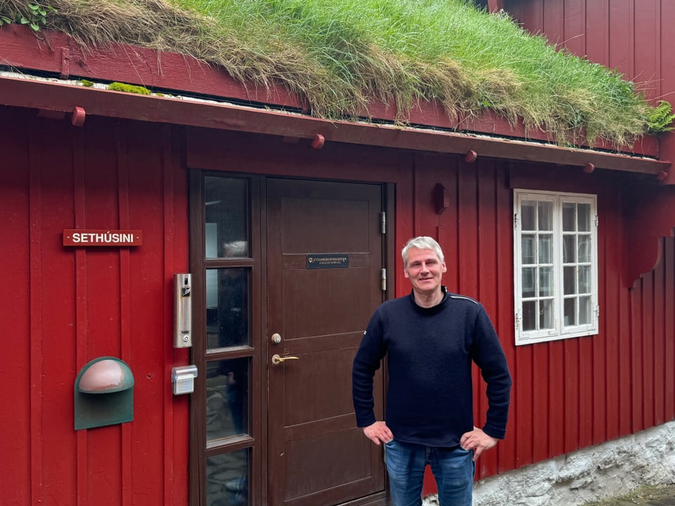 Mann vor rotem Holzhaus mit Grasdach.