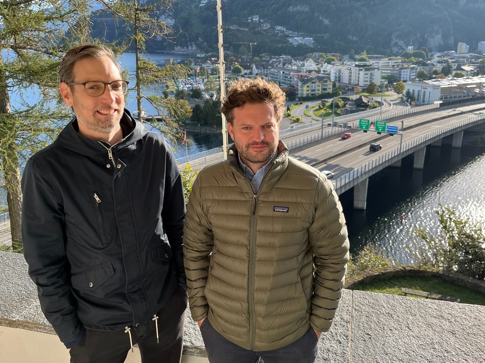 Zwei Männer stehen draussen mit einer Brücke und Stadt im Hintergrund.