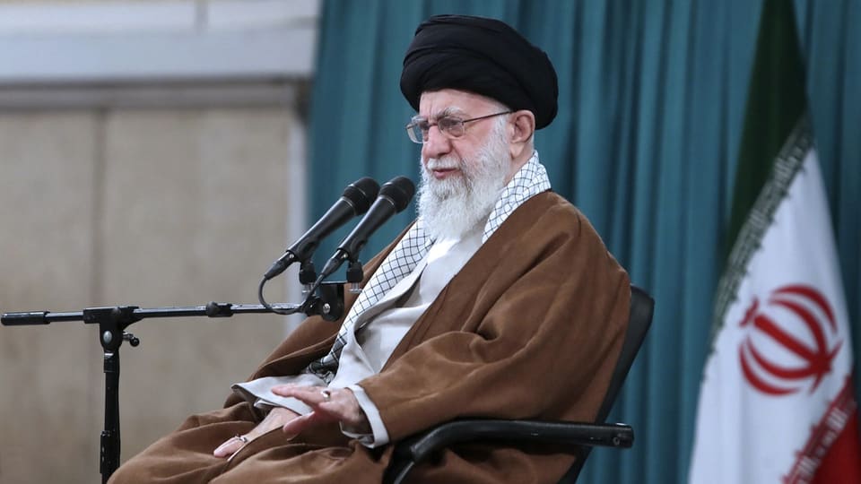 Älterer Mann mit Bart in brauner Robe spricht in Mikrofon, iranische Flagge im Hintergrund.