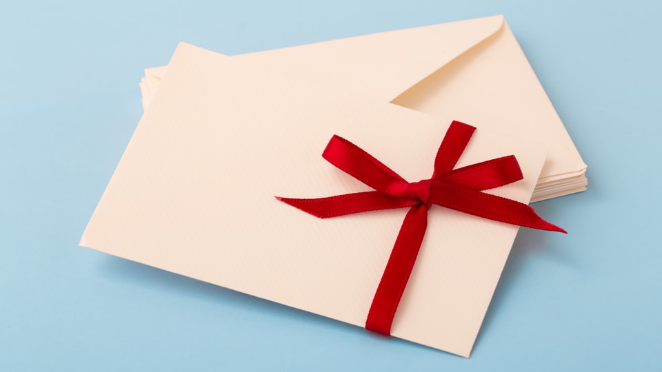 Ein Briefumschlag mit roter Schleife vor blauem Hintergrund.