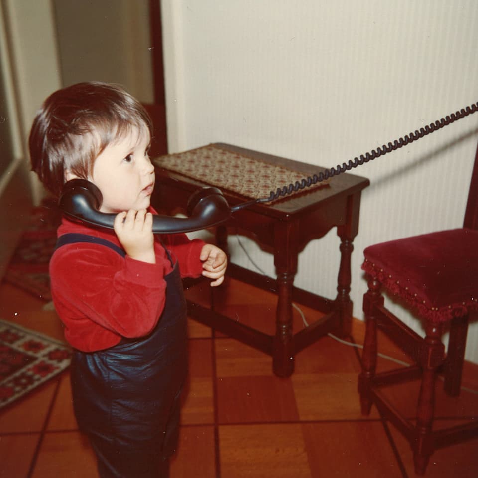 Ein kleiner Bub mit rotem Pullover und blauer Latzhose hält sich einen grossen schwarzen Telefonhörer ans Ohr.