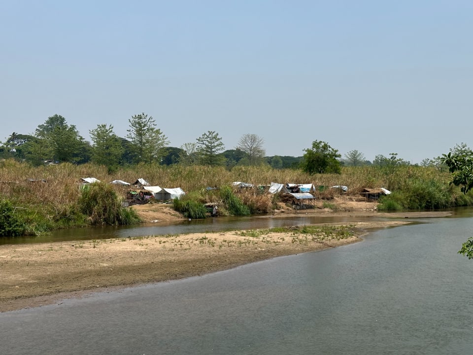 Flussufer mit rudimentären Hütten und Vegetation im Hintergrund.