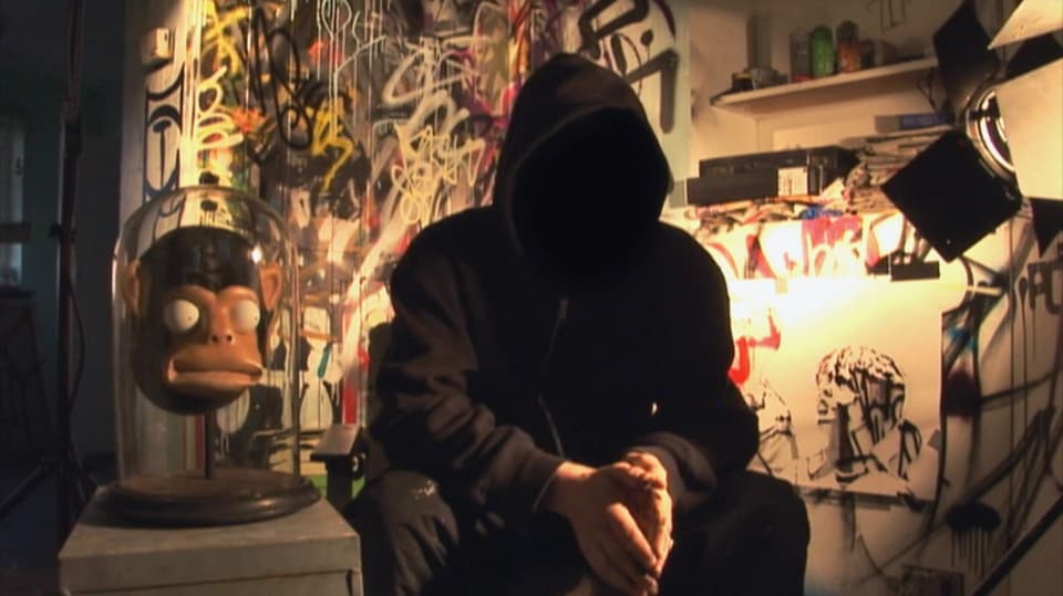 Eine dunkel gekleidete Person mit Kapuzenpulli im Scheinwerfer eines Lichts. Im Hintergrund diverse Streetart-Objekte