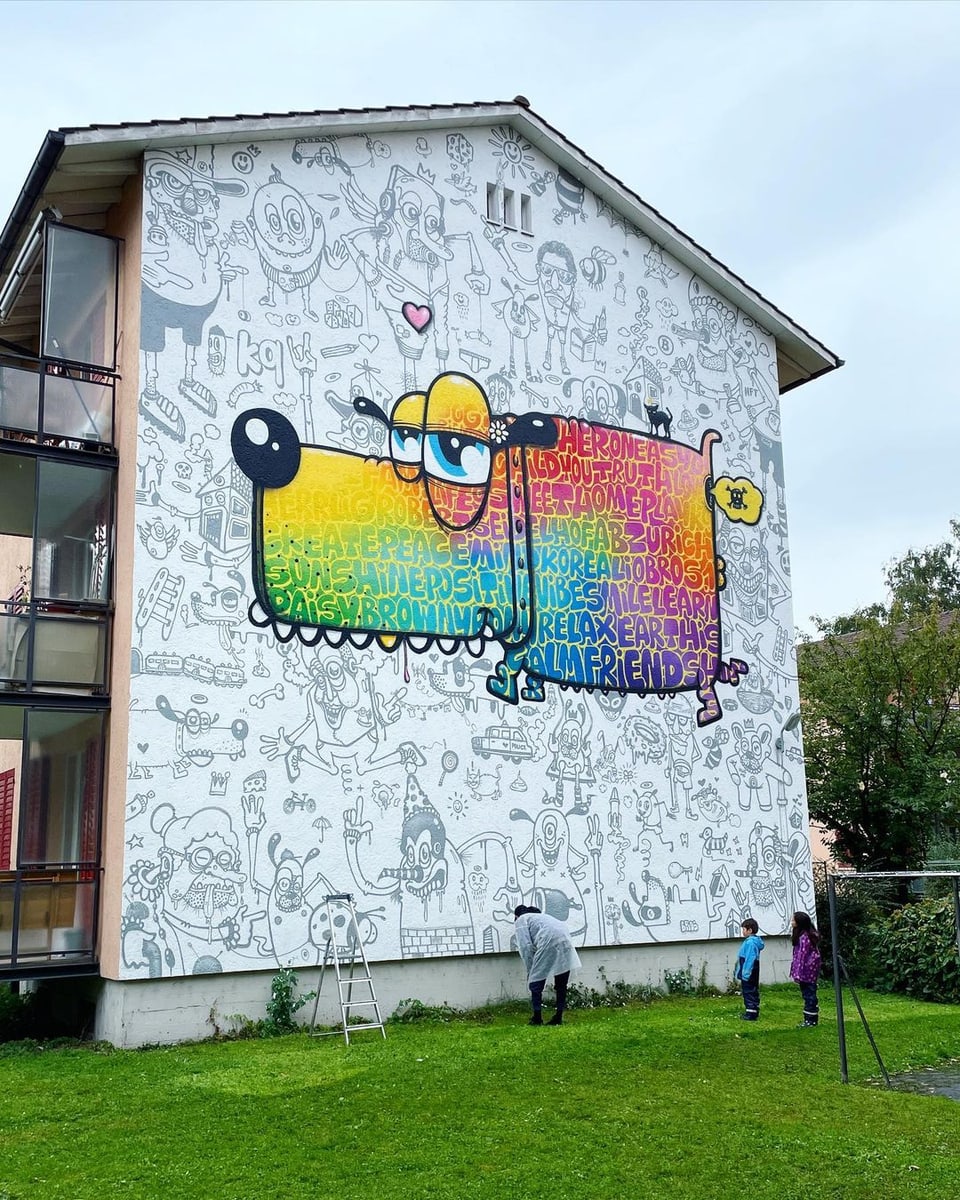 Ein Graffiti-Künstler finalisiert das Design einer verzierten Hausfassade. Zu sehen ist ein grosser farbiger Hund.