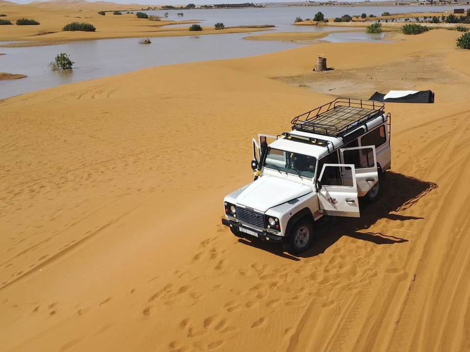 Geländewagen in der Wüste mit offener Tür, Sanddünen und Wasserstellen im Hintergrund.