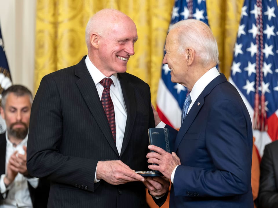 Zwei Männer schütteln sich die Hände und lächeln, einer hält eine Medaille. Amerikanische Flaggen im Hintergrung.