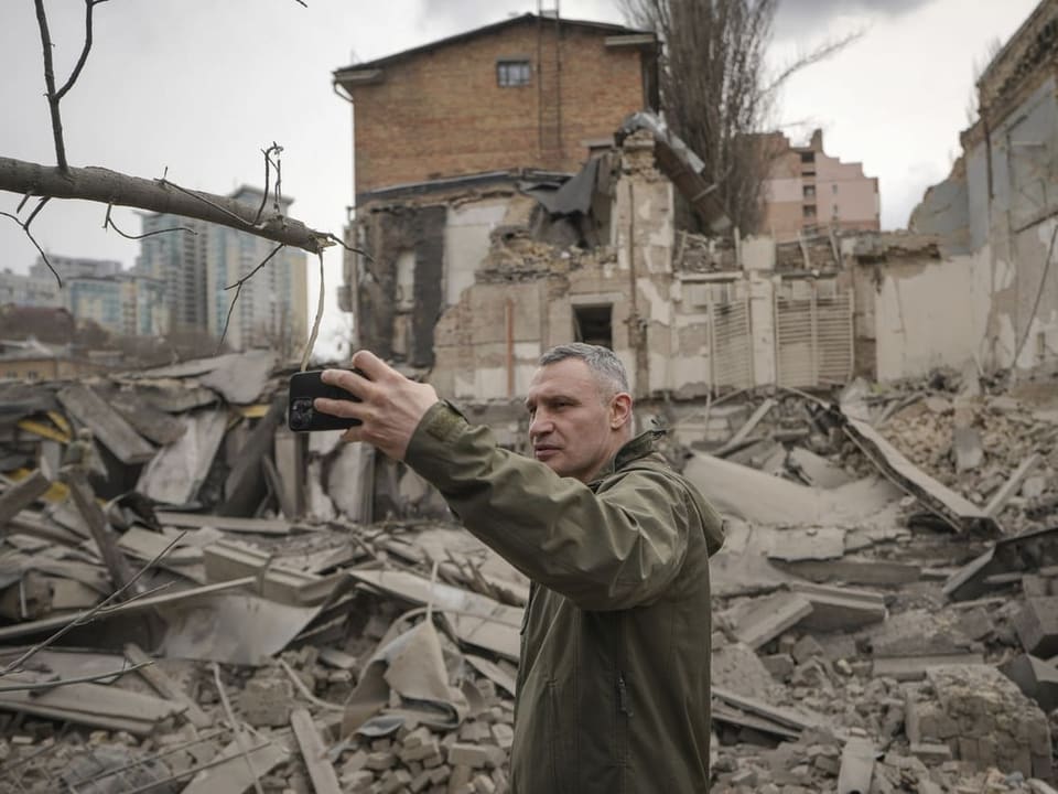Mann vor zerstörten Gebäuden. Er hält das Handy vor dem Gesicht.