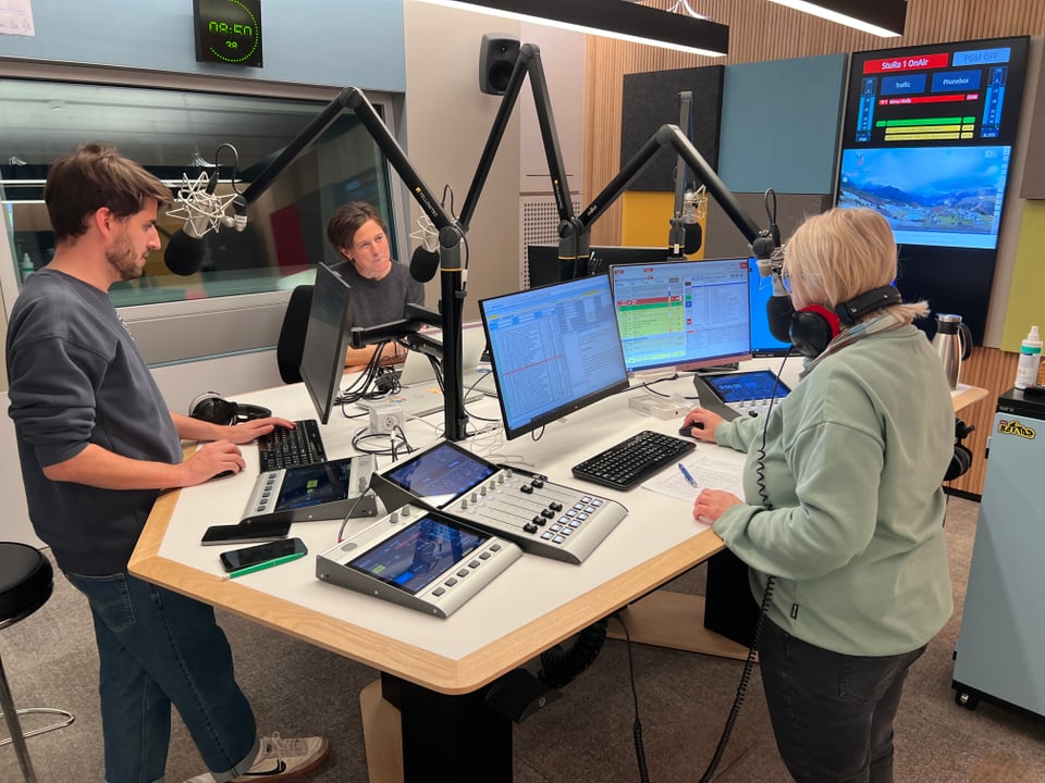 Drei Personen arbeiten in einem Radiostudio mit Mikrofonen und Monitoren.