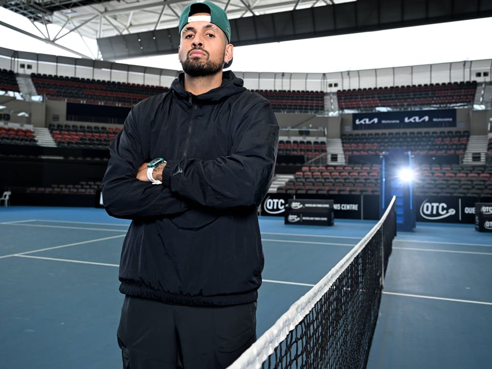 Person auf Tennisplatz mit gekreuzten Armen vor Netz in leerem Stadion.