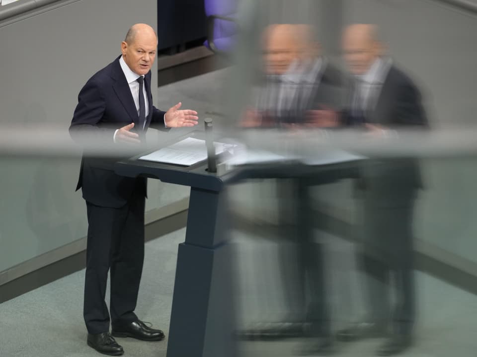 Mann in Anzug spricht an einem Podium, Spiegelbild sichtbar.