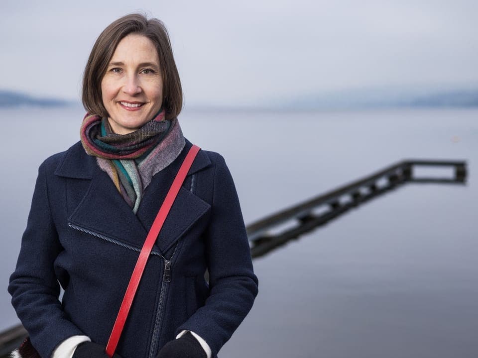 Frau mit Schal steht vor einem See und einem Steg.