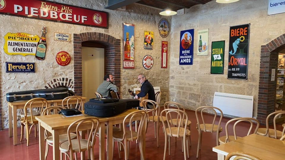 Innenraum eines Cafés mit zwei Männern an einem Tisch, Retro-Werbeschilder an der Wand.