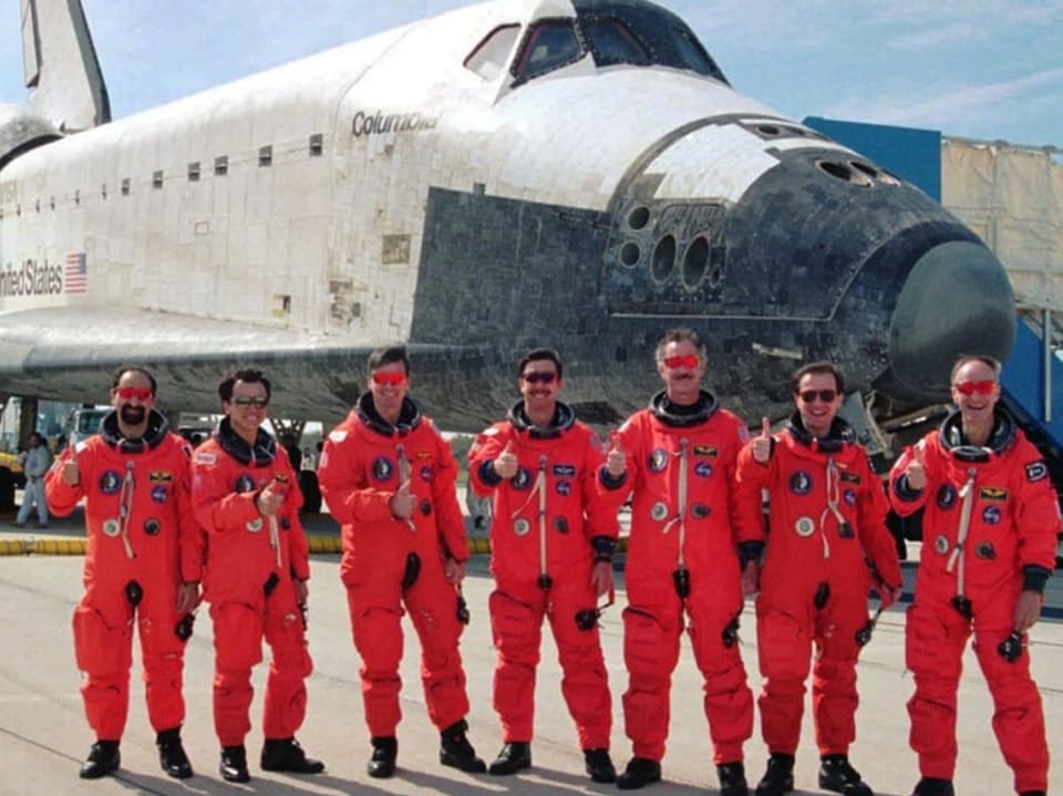 Eine Gruppe von sieben Astronauten in orangenen Raumanzügen vor einem Space Shuttle.