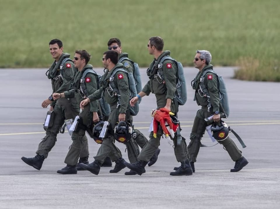 Sechs Piloten in grüner Uniform marschieren über das Flugfeld.