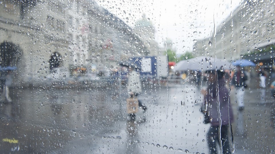 Regentropfen rinnen über eine Scheibe. Im Hintergrund ziehen Menschen mit Regenschirmen vorüber.