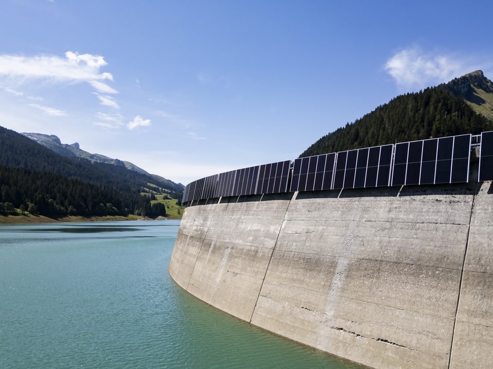 Staumauer mit Solarpaneelen vor Berglandschaft.