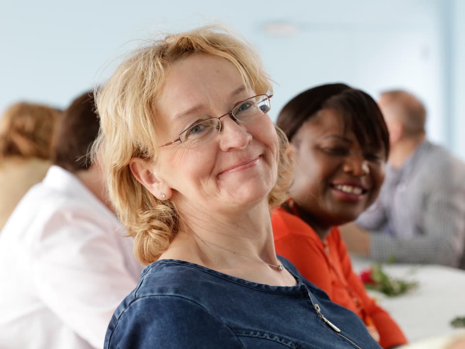 Eine blonde Frau sitzt am Tisch und lächelt.