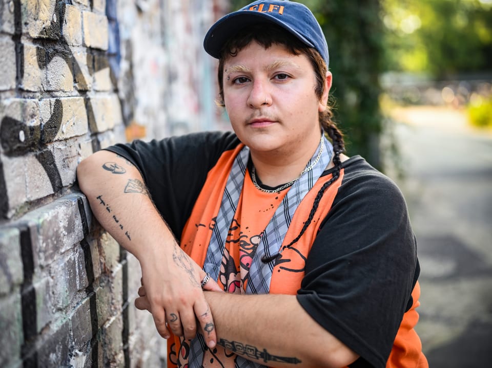 Person mit Tattoos lehnt an einer Backsteinmauer.