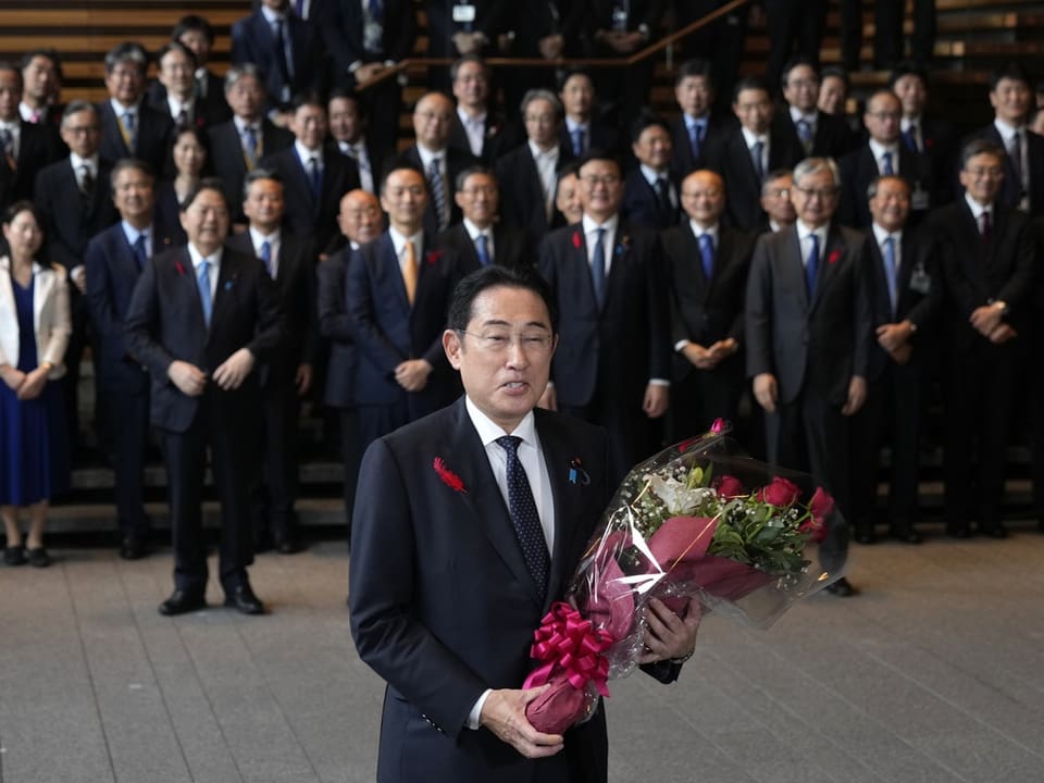 Fumio Kishida hält Blumenstrauss vor Gruppe von Menschen in Anzügen.