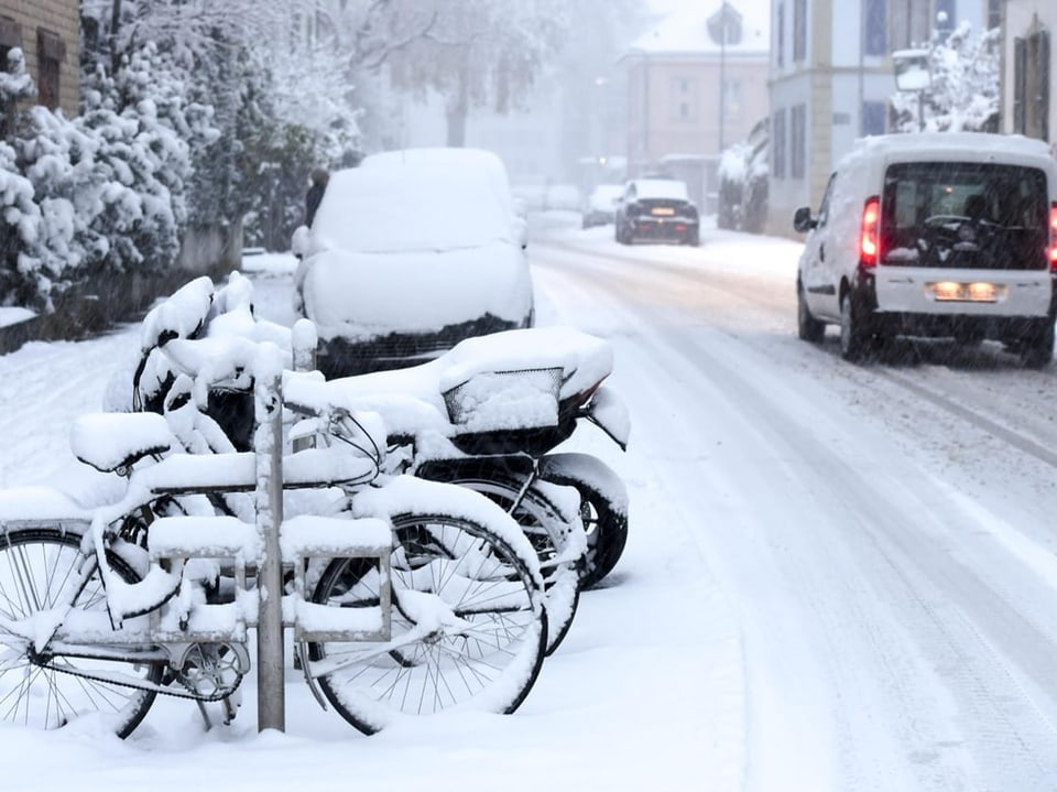 Verschneite Strasse mit parkenden Fahrrädern und Autos.