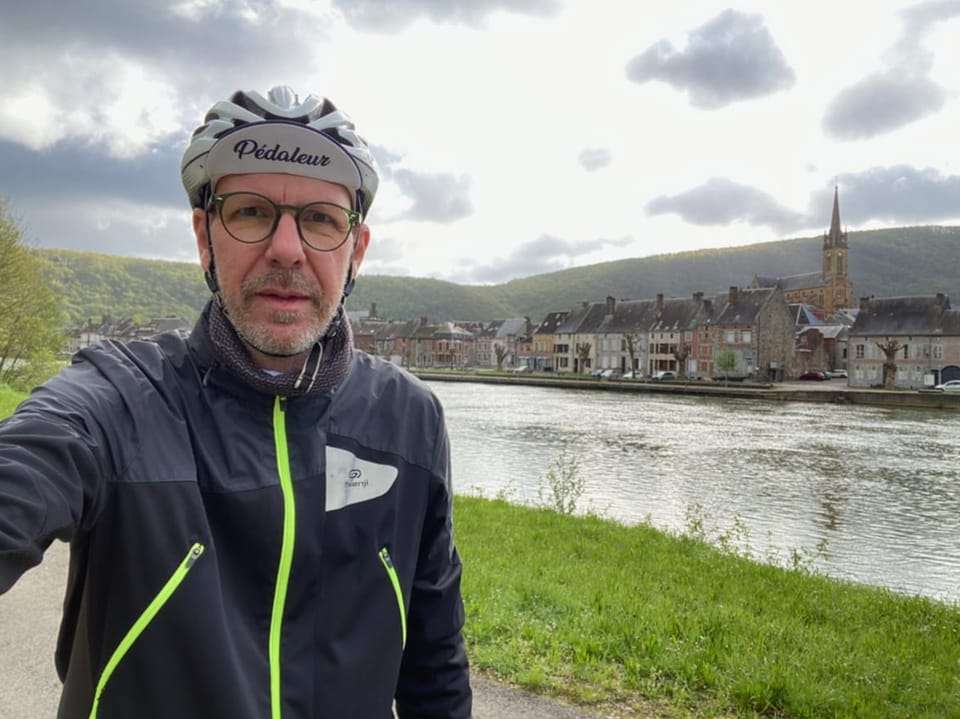 Radfahrer macht Selfie vor Fluss und Dorf.